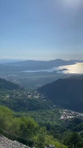 A vertical video shot from a high vantage point looking down on a bay surrounded by mountains. The sky is clear blue with a bright sun creating a glare in the upper right corner. The sun reflects off the water in the bay, creating a shimmering path of light. Lush green trees cover the mountainsides, and several small villages with red-tiled roofs are nestled in the valleys and on the slopes. A road winds through the landscape, visible in the middle ground. In the foreground, there is a rocky, gravelly area with some green foliage and a few trees. The video appears to be taken on a sunny day.