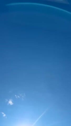 The video shows a bright sun in a clear blue sky with a few wispy clouds. Lens flares are prominent, creating circular and linear patterns of light. The sun is positioned in the lower portion of the frame, shining intensely and emitting rays of light. A faint, elongated cloud is visible below the sun, and two small, fluffy clouds are to the left of the sun. The overall environment appears to be outdoors on a sunny day.