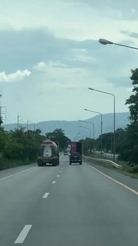 The video shows a view from a vehicle driving on a highway. There are three trucks visible in front of the camera vehicle. The truck closest to the camera is a cement mixer truck. Behind it is a black pickup truck. Further ahead is a larger red and white truck. The highway is paved with asphalt and has white lane markings. On the right side of the road, there is a yellow line marking the edge of the lane, followed by a guardrail and then vegetation. Along the side of the highway, there are many streetlights. In the distance, there are trees and mountains under a cloudy sky. The overall scene depicts a typical highway commute on a cloudy day.