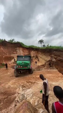 A green dump truck is shown in a dirt quarry. The truck is moving. There are several people visible in the quarry, some are standing and watching the truck, others appear to be working. The environment is rough and uneven with piles of dirt and rocks. The sky is cloudy.