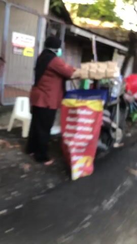 The video captures a bustling street scene in Candi, East Java, likely during the daytime given the bright lighting. The camera, moving briskly, pans across a street where vendors are preparing to sell their goods.

In the foreground, a person wearing a face mask and a reddish-brown top is attending to a cart filled with what appears to be snacks or food items, wrapped in light-colored packaging. The cart itself is adorned with a vibrant red and yellow banner, though the text on it is blurred due to motion. Adjacent to this cart, a motorcycle is parked, equipped with a rack holding more packaged goods.

The scene then shifts to show another vendor, this time a woman in a pink headscarf and a green top, interacting with a bicycle-powered cart. This cart also carries numerous items, some of which seem to be food products. Behind her, a man is visible near a storefront, and further down the street, another motorcycle is parked, carrying a stack of what could be eggs or similar small items. The overall impression is one of a market or street-side commerce.

The ground appears wet in places, suggesting recent rain or ongoing cleaning. The presence of multiple vendors, motorcycles, and the general activity implies a lively commercial area. There's a sign with "PMAKHISMA INDONESIA" visible on a building in the background. The fast-paced, somewhat shaky nature of the video conveys a sense of movement and the energetic atmosphere of the location.