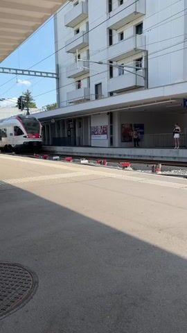 A train is stopped at a station. The train is white and red with blue accents. A woman wearing a white top and black skirt stands on the platform. A man in a blue shirt and dark pants is also on the platform. The station has a covered waiting area and a tall white building behind it. The sun is shining and the sky is blue.