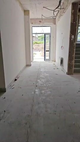The video shows an interior hallway of an unfinished building. The walls are white and appear to be partially plastered. The ceiling is exposed, with electrical wiring and conduit visible. The floor is tiled with large, light-colored tiles that show signs of dust and debris. A doorway at the end of the hallway leads outside. Through the doorway, a person is visible working in an outdoor area. The outdoor area appears to be a construction site, with uneven ground, some piles of materials, and buildings in the background. A metal radiator or vent is mounted on the right wall. Various pieces of debris, including foam insulation and possibly plaster scraps, are scattered on the floor. The lighting is overcast, suggesting a cloudy day.