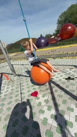 A young boy is sitting on an orange ball swing in a playground on a sunny day. He is wearing a white t-shirt and gray shorts and is barefoot. He is holding onto a blue rope attached to the ball. The playground has a patterned tile floor and is surrounded by colorful dome-shaped structures. A red hat and a pair of sandals are on the ground nearby. A person's legs and shadow are visible in the foreground, suggesting they are filming the boy. The camera angle shifts slightly during the short video.