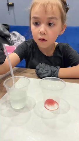 A young girl wearing black gloves is carefully adding a clear liquid from a pipette into a plastic cup. Beside it, another plastic cup contains a small amount of red liquid. The girl is focused on her task, her eyes looking down at the cups. She is seated at a table, with a blue couch visible in the background. The scene suggests a science experiment or a craft activity.