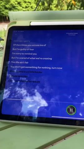 This is a close-up shot of a tablet displaying lyrics and a music player interface. The tablet is resting on a green outdoor table, with a blurred background of lush green foliage, suggesting an outdoor setting, possibly a garden or park. The time on the tablet's display indicates 17:10, and the date is "Чт 2 июля," which translates to "Thursday, July 2nd." The song title is "It Could Be Sweet" by Portishead. The lyrics displayed on the screen are:

"Of the crimes you accuse me of
But I'm guilty of fear
I'm sorry to remind you
But I'm scared of what we're creating
This life ain't fair
You don't get something for nothing, turn now
Mmm, gotta try a little harder
It could be sweet
Like a long forgotten dream
And we don't need them"

A music player control bar is visible at the bottom of the screen, showing the current playback time at 0:51 and the song duration or end time at 0:51. A green circular button with a pause symbol (two vertical bars) is prominently displayed on the right side of the player interface, indicating the music is currently playing or was paused. The screen is reflective, showing a partial reflection of the sky and greenery. The overall mood conveyed by the image is one of quiet contemplation, with the music and lyrics set against a natural backdrop. The weather appears to be pleasant, likely sunny given the brightness of the foliage. While no people are directly visible, the presence of the tablet implies someone is interacting with it. The setting feels serene and perhaps a bit melancholic, fitting with the song's title and lyrics.