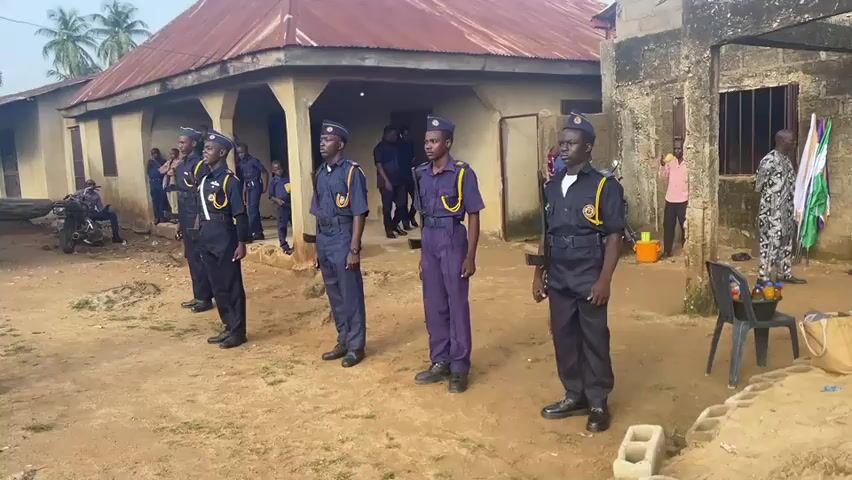 The video shows a group of at least six young men in matching blue and black uniforms with yellow trim, standing in formation in an outdoor courtyard. They appear to be part of an organization or a youth group. The men are standing at attention, facing the camera. In the background, there is a building with a red, corrugated metal roof and light-colored walls. To the right of the building, there are a few people standing around, and a flag with green and white colors is visible. The ground is dirt and gravel. The overall atmosphere appears to be formal, possibly a ceremony or a gathering.