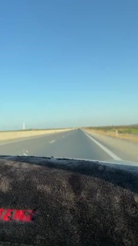 The video shows a view from inside a car driving on a highway. The road is clear with a few other vehicles visible in the distance. The sky is a clear blue and the weather appears to be sunny. The dashboard of the car is visible in the foreground, covered with a dark fabric.