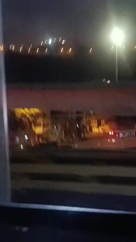 The video shows a night scene from what appears to be a moving vehicle, likely a train or bus. Outside, one can see blurry city lights in the background, and some buildings visible through a window or opening. The view is somewhat obstructed by parts of the vehicle, especially the red-colored top frame. The lighting conditions are dim, typical of a night scene with artificial lights.