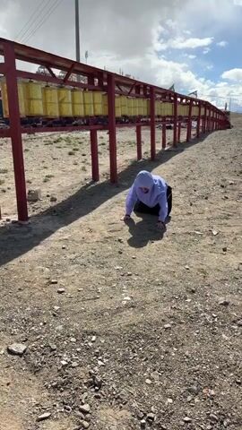 In an outdoor, arid setting, a person dressed in a light purple hooded jacket and black pants is performing repeated full-body prostrations on a gravelly dirt ground. The individual extends their body forward, lies flat on the ground, brings their hands together in a prayer-like gesture, and then rises to repeat the motion.

Behind the person, a long row of large, golden prayer wheels is visible, mounted on a red metal structure. The ground is dry and covered with small stones and dirt. In the background, under a partly cloudy sky, some utility poles or structures can be seen in the distance. The sun is shining, casting distinct shadows on the ground. An audible voice can be heard counting in what sounds like Russian. The scene suggests a devotional or religious practice, likely at a Buddhist site given the presence of prayer wheels.
