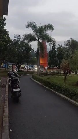 The video shows an outdoor scene. It appears to be near a roadside with trees, grassy areas, and a walkway or small road. There's a large, stylized monument in the background, colored in shades of red, orange, and yellow. The sky is overcast, suggesting it may be a cloudy day. A scooter is parked along the roadside at the beginning of the video. There appears to be a highway with cars in the background of the video. A call to prayer sounds are heard during the recording.