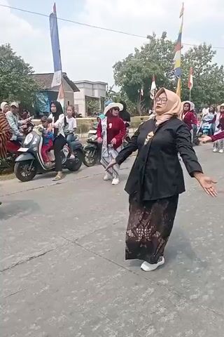 This video shows a group of people participating in an outdoor procession or parade. In the foreground, a woman in a black top, a beige headscarf, and a patterned skirt is dancing with her arms outstretched. Behind her, another woman in a red jacket, a white hat, and a striped skirt is also dancing. To the left, a man in a striped shirt and sarong is also dancing. Several motorcycles are parked along the side of the road, and spectators are visible in the background. The environment appears to be a street with buildings and trees lining the side. The sky is bright, suggesting it is daytime. There are flags and banners visible, indicating a celebratory event.