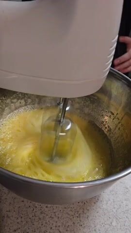 The video shows a close-up of a white electric hand mixer with silver beaters being used in a stainless steel bowl filled with a yellow, frothy liquid, likely eggs. The mixer is in motion, causing the liquid to churn and create bubbles. The bowl sits on a speckled gray countertop. A person's hands are partially visible, holding the mixer. The lighting is bright, and the background is blurred, suggesting a focus on the action in the bowl. The overall impression is of a cooking or baking process.