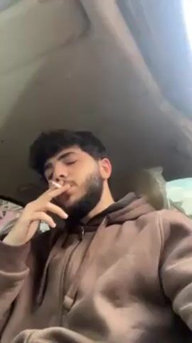 A young man with dark curly hair and a beard is sitting in the driver's seat of a car, wearing a brown hoodie. He is smoking a cigarette, exhaling smoke rings that float towards the car's ceiling. The interior of the car is visible, with the roof and part of the windshield in the frame. The car seat is covered in plastic. The lighting is bright, suggesting it's daytime.