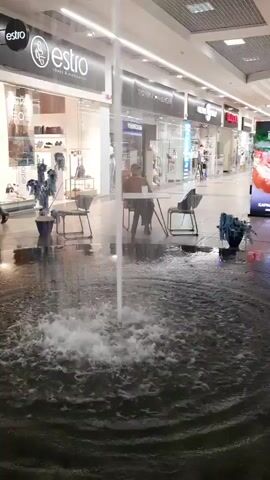 The video shows an indoor scene in what appears to be a shopping mall. In the foreground, a fountain is active, with water spraying upwards and splashing into a basin. The water spray is a prominent visual element, creating a blurred effect in the center of the frame. In the mid-ground, there are several chairs and a small table near the fountain. A person is seated at the table, facing away from the camera. Behind the fountain and seating area, a walkway stretches into the distance, lined with various storefronts. Signs for "estro" and "tommy hilfiger" are visible on the storefronts. The mall appears to have multiple levels, with a railing and another walkway visible on an upper level. The lighting is typical of an indoor commercial space, with overhead fluorescent lights illuminating the area. The overall impression is of a public indoor space with a decorative fountain feature.
