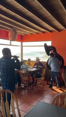 A video depicts a restaurant scene with musicians performing for diners. In the foreground, a man in a plaid shirt holds a microphone and sings. To the right, another man plays a tuba. Two diners are seated at a table in the middle ground, one wearing a cowboy hat. The restaurant has an orange wall and a wooden ceiling, with a view of the ocean through a large window.