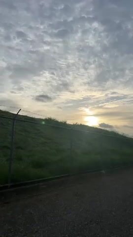 The video shows a sunrise over a grassy hill with a chain-link fence in the foreground. The sky is cloudy, with the sun breaking through the clouds and casting a bright light. The grass on the hill appears to be wet, possibly from dew or recent rain. A paved path runs along the bottom of the frame.