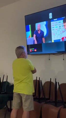 A man in a bright yellow t-shirt and khaki shorts stands with his back to the camera, watching a large television screen. He has his arms crossed and appears to be focused on the broadcast. The TV displays a sports broadcast, likely soccer, with two men on screen. One man is seated on a blue couch, wearing a dark blue polo shirt, while the other man is on the right side of the screen, wearing a white and red jersey. Below the men on the screen, a scoreboard shows the score "0-1". The scene takes place indoors, with several chairs stacked behind the man. The lighting suggests it is daytime or a well-lit interior. The man's posture and focus imply he is engaged in watching the game. Visible text on the TV screen includes "FIFA", "QATAR", "VISA", and various other sponsor logos. The setting appears to be a public space or a room equipped for watching sports.