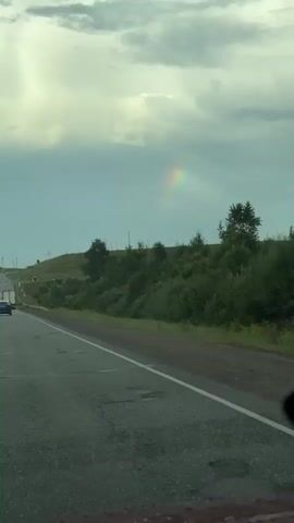 The video shows a view from a moving vehicle on a paved road. The road is grey and has some potholes. On the right side of the road, there is a grassy embankment with trees and bushes. In the distance, on the left side of the road, there is a green hilly landscape. The sky is cloudy and grey with some lighter patches. A faint rainbow is visible in the sky above the trees. There is a car in the distance on the left side of the road. The camera appears to be looking out the windshield or a side window of the vehicle.