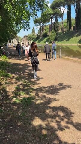 The video shows a sunny outdoor scene with a path alongside a body of water. A red-haired woman in the foreground is walking towards the camera, looking at her phone, and then smiles at the camera. She is wearing a black shirt, gray pants, and white sneakers, with a black jacket slung over her shoulder and a white bag. Other people, some walking and some standing, are visible on the path behind her. On one side of the path are trees and the ruins of a building, while on the other side is a grassy bank and a line of trees. Shadows from trees are cast onto the path. The sky is blue with some clouds.