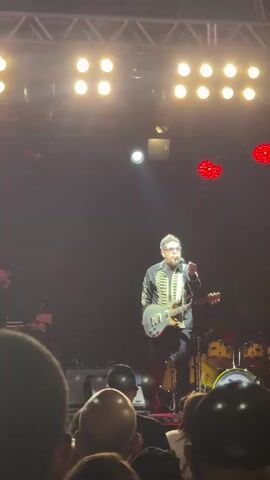 A man in a black jacket with gold trim and a guitar is on stage performing. He is wearing glasses and has a beard. There are lights above him and a band behind him playing drums and other instruments. A crowd of people is in the foreground.