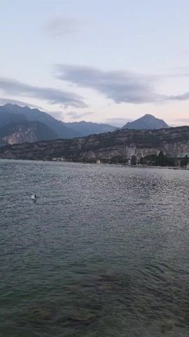 This is a video taken from a moving drone or camera, panning across a body of water towards a distant shoreline. In the foreground, the water is clear enough to see rocks and the seabed beneath the surface. There is a small white object, possibly a bird or debris, in the water to the left of the frame. The shoreline features a range of mountains in the background, with a town or village built along the water's edge. Buildings with colorful roofs are visible, and there appears to be a small pier extending into the water. The sky is overcast with scattered clouds, suggesting either early morning or late evening. The overall impression is a serene landscape.