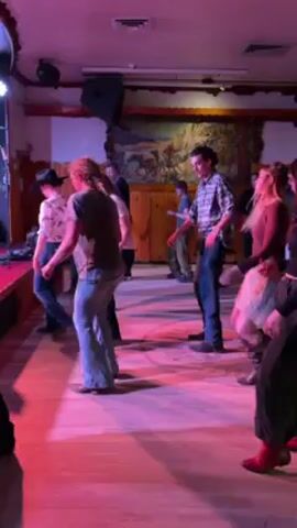 The video shows a group of people line dancing in a room with a decorated wall. The room is lit with pink and blue lights. The dancers are wearing a variety of casual clothing, and at least one dancer is wearing a cowboy hat. The camera holder appears to be standing in the middle of the crowd.