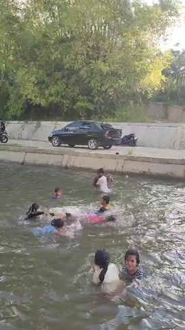 This media file shows a group of children swimming and playing in a body of water, likely a river or canal, in Saweuk, Indonesia. The water is murky and appears to be relatively shallow, with several people submerged up to their shoulders or chests. In the foreground, two young girls are visible, one with long dark hair and the other with shorter dark hair, both looking towards the camera with smiles. Behind them, more children are scattered throughout the water, some splashing and others floating on inflatable toys. The general mood appears cheerful and playful.

The background reveals a concrete embankment with a black sedan parked on it. Beyond the embankment, lush green trees and foliage dominate the landscape, suggesting a natural or semi-natural environment. The lighting indicates it is daytime, possibly late afternoon, with sunlight filtering through the trees and casting a warm glow. There is no visible text in the media file. The scene captures a moment of simple recreation and enjoyment among the children in a tropical setting.