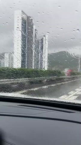 The video shows a view from inside a vehicle driving on a wet road during a rainstorm. The windshield is covered in raindrops, distorting the view of the outside. In the distance, there are tall buildings and a highway with other vehicles. The road is slick with water, and the sky is overcast, indicating heavy rain. The perspective suggests the video is recorded by a passenger or driver.