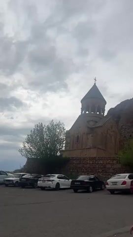 A video pans from left to right, revealing a historic stone church with a conical dome and cross on the left under a cloudy sky. A gravel parking area in the foreground and midground contains several parked cars of various colors, including white, black, red, silver, and dark blue. Behind the cars and to the right of the church, a large stone archway stands, leading into an open area. Rocky, barren mountains rise steeply behind the archway and church. Several people are visible walking near and through the archway. Green foliage is scattered around the base of the church and near the archway.