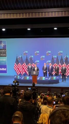 A man in a dark suit and a tie stands at a podium with the presidential seal on it, speaking to a crowd of reporters. He is on a stage in front of a blue backdrop with NATO logos and American flags. He is flanked by several other men in suits, some of whom are standing behind him. The reporter's cameras are visible in the foreground, some are set up on tripods.