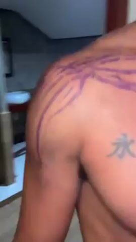 A shirtless man stands indoors, showing a purple, sketchy design on his right shoulder. He also has a tattoo on his upper chest written in Asian characters. A doorway is partially visible behind him, and a peek into another room is seen through it. Music is playing in the background.