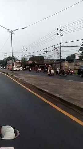 The video shows a view from a motorcycle as it travels along a paved road. Several other motorcycles and a bus are also visible on the road. The bus is red and white with an orange stripe. There are buildings and trees lining the road, as well as utility poles and wires overhead. The sky is overcast. A person wearing a black shirt and a black helmet is riding the motorcycle in the foreground, with a black backpack visible on their back. The license plate of the motorcycle is visible and reads "L 3451 PU". Another motorcycle is visible to the right of the bus, carrying two people. A red crane is visible in the background behind some buildings.