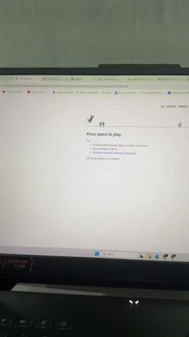 The video shows a laptop screen displaying a Chrome browser window. The browser is displaying a "No internet" error page, featuring the familiar Chrome dinosaur game. The dinosaur is shown with cacti and other obstacles in its path, indicating the game is in progress. The text "Press space to play" is visible below the dinosaur, and a list of troubleshooting steps is also displayed. The bottom portion of the screen shows a keyboard and a portion of the laptop's casing, which appears to be black with some sort of decorative detailing. The video captures several brief moments, showing the dinosaur moving across the screen, and then the text "GAME OVER" appearing.