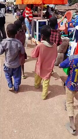 The video shows a group of young children walking down a crowded street lined with market stalls. The camera follows them from behind as they move along the pavement. The children are dressed in casual clothing, with one wearing a jersey with the name "Jackson" and the number "15" on the back. The street is busy with people and stalls selling various goods. The stalls are covered by colorful umbrellas. Some of the people in the background are browsing the merchandise, while others are simply passing through. The overall atmosphere is lively and bustling, characteristic of a busy marketplace.