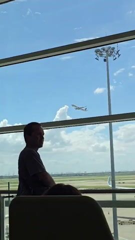 A man stands by a large window in an airport terminal, looking out at the tarmac and a clear blue sky with scattered clouds. An airplane is visible in the distance, ascending into the sky. A tall light pole with multiple lights stands outside the window. In the foreground, two chairs are visible, one occupied by a person with a pink neck pillow. Another person's head is visible in the lower center of the frame, also seated. The scene suggests a moment of quiet observation within the busy environment of an airport.