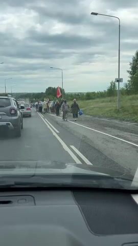 A group of people are walking in a procession along the side of a road. The sky is overcast with clouds. The procession is moving from left to right in the frame. There are cars on the road, some of which appear to be stopped or moving slowly. The people in the procession are dressed in a variety of clothing, and some are carrying flags. The procession is on the right lane of the road, and the cars are on the left lane. The road has two white lines marking the lanes. There is a ditch and vegetation on the right side of the road. A lamppost is visible on the right side of the road. The video is shot from inside a vehicle, likely from the dashboard.