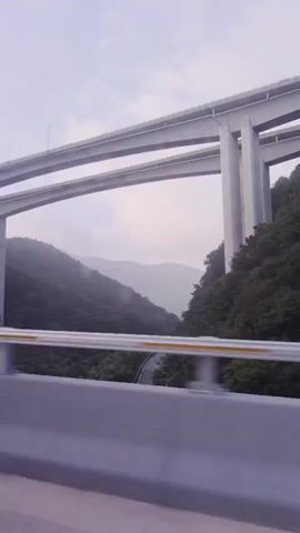 This is a vertical, low-angle, exterior shot of a multi-tiered highway bridge passing over a lush green mountain valley. The bridge is made of concrete pillars and sleek, modern roadways. The lower road is visible, winding through the valley. The sky is overcast, creating a soft, diffused light. Trees cover the slopes of the mountains, and a few distant houses are visible in the valley. The camera is positioned from a vehicle traveling on a road parallel to the bridge, with a gray barrier and a yellow stripe in the foreground.