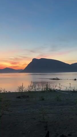 A serene sunrise paints the sky with vibrant hues of orange, pink, and blue over a calm body of water. Silhouetted mountains rise in the distance, their shapes softened by the morning mist. The foreground features a dark, sandy shore dotted with sparse vegetation. The video pans slowly, offering a panoramic view of the tranquil landscape. The water reflects the colorful sky, creating a mirror-like effect that enhances the natural beauty of the scene.