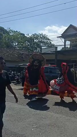 The video captures a street scene featuring performers in elaborate costumes. In the foreground, a person in black clothing raises their hand, palm facing the camera, in a gesture that could be interpreted as a stop or greeting. To the right, a performer in a vibrant red and black costume with a large, circular headdress adorned with sequins and a crest is visible. Further to the right and slightly behind, another performer in a similar red and black costume with layered, ruffled fabric is seen. To the left, several other performers in colorful, detailed costumes, including a figure with a straw hat and intricate facial markings, are present. Some of these performers appear to be engaged in a dance or procession. In the background, a dark-colored vehicle and other onlookers are visible, along with buildings and trees under a bright, sunny sky. The ground appears to be asphalt. The overall atmosphere suggests a cultural event or performance taking place outdoors.