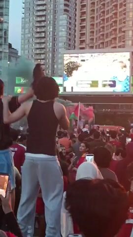 A crowd of people are gathered outdoors, watching a large screen that displays a sporting event. Many in the crowd are wearing red shirts, some with the name "Ronaldo" and the number "7" on the back. A Portuguese flag is visible, waving in the air, and green and pink smoke is being released. People are holding up their phones, recording the event. The background consists of tall buildings. The time stamp indicates the event is occurring at dusk.