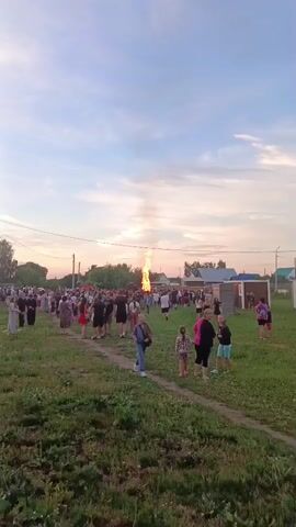 A large crowd of people are gathered in a grassy field, watching a tall bonfire blaze in the distance. The sky is partly cloudy and the sun appears to be setting, casting a warm glow on the scene. The people are casually dressed, and many appear to be families with children. Some are standing in groups, while others are walking through the field. The bonfire is the central focus of the gathering, and the flames are reaching high into the sky. The environment appears to be a rural area, with houses visible in the background. The overall atmosphere seems festive and communal.