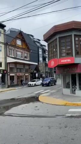 The video shows an urban street scene with a variety of buildings, cars, and pedestrians. The buildings are of different architectural styles, ranging from traditional to modern. Several businesses such as a hostel, a clothing store called "Urbanmix", and various other shops are visible. Cars are seen driving on the road. Pedestrians are walking along the sidewalks. The weather appears cloudy and overcast.