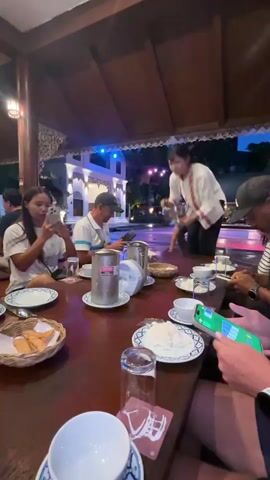 A group of people are seated around a wooden table at an outdoor restaurant. The table is set with plates of food, rice, and drinks. A server in a white uniform is attending to the table, and some of the diners are using their phones. The background shows a building with colorful lights and some greenery. The lighting is dim and appears to be evening.