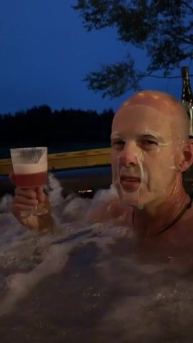 A man with a face mask on is in a hot tub with foamy water. He is holding a glass with a pink drink in it. The time is evening, and the sky is dark blue. There are trees in the background. A sign is visible on a wooden post next to the hot tub. The man turns his head to look at the camera.
