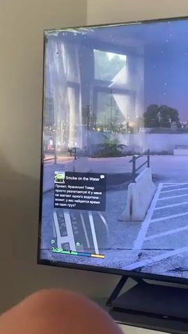 The video shows a person playing a video game, Grand Theft Auto V, on a large screen television. The game displays a nighttime scene with a character standing in a parking lot. The character is wearing a dark vest over a long-sleeved shirt and appears to be looking away from the camera. The parking lot is dimly lit by streetlights and has a concrete barrier in the foreground. A mini-map is visible in the bottom left corner of the screen, indicating the character's location within the game world.

A pop-up message in the game, in Russian, reads: "Hi, Franklin! The tow truck is coming, and I don't have any drivers - maybe we'll find time for one more cargo!"

The time of day in the game appears to be evening or night, with a starry sky. The setting is urban, with buildings and palm trees visible in the background. The television is placed on a stand, with a game controller resting on it. A license plate with Cyrillic text, "КРИВБАСС", is visible on the stand below the television. The overall mood seems casual, as the person is engrossed in playing the video game. The location context of Vejprty, Czechia, is provided but not visually evident within the game itself.