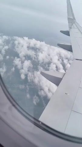 The video shows an aerial view from an airplane window. A portion of the airplane's wing is visible on the right side of the frame, extending towards the top of the image. Below the wing, a vast expanse of white, fluffy clouds fills the middle ground. Patches of land with some urban development are visible beneath the clouds. The sky is overcast and appears to be a muted grey. The camera perspective suggests the plane is in flight, possibly ascending or descending, as indicated by the dynamic cloud formations. The lighting is soft and diffused due to the cloudy conditions.