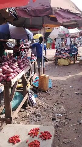 The video shows a bustling outdoor market, likely in an African country. Several vendors have set up shop under umbrellas, displaying their goods on tables. There are piles of red onions on one table, and what appear to be red chili peppers are laid out on a small table in the foreground. A man wearing a blue shirt and shorts stands near the onions, while several other people mill about, some examining the merchandise. Across from the onion stand is a table with clothing for sale, and a woman sits on a bench nearby. The ground is a mix of dirt, gravel, and scattered debris, giving the area a somewhat unkempt appearance.