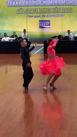 A young boy and girl are dancing a ballroom dance routine on a polished wooden floor. The boy is wearing a black, long-sleeved shirt and black pants. The girl is wearing a bright pink, sleeveless dress with a full skirt and a matching headband. They are facing each other, with the boy on the left and the girl on the right. The girl's left arm is raised above her head, and her right arm is extended to the side. The boy's arms are extended to his sides, and he is looking at the girl. In the background, a banner reads "CUP GRACE DANCE NAM 2026". Several people are seated at tables in the background. The lighting is bright, and the scene appears to be an indoor event.
