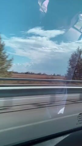 The video shows a view from inside a vehicle, looking out a window at a roadside landscape. The scene is in motion, indicated by the blur of trees and the roadside barrier. The sky is blue with scattered white clouds. In the foreground, a road barrier with reflective white and gray elements is visible. Beyond the barrier, there are fields and trees. The reflection of the interior of the vehicle, including what appears to be a metallic object, is visible on the window. The time stamp progresses from 00:00 to 00:04, suggesting a short duration of recording.